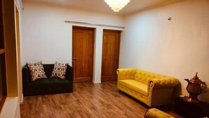 Una habitación con un sofá verde, un sofá amarillo y suelo de madera. Hay dos puertas de madera y una araña.