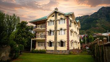 A multi-story building with a balcony on the second floor, surrounded by greenery and mountains in the background.