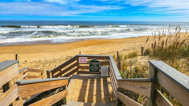 Un escalier en bois mène à une plage où un panneau indique : « Il est interdit de marcher sur les dunes » et « Des tentes et des parasols sont disponibles à la location ».