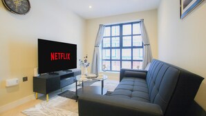 Ein Wohnzimmer mit einem großen Flachbildfernseher, der 'NETFLIX' anzeigt, einem dunkelgrauen Sofa, einem kleinen Couchtisch und einem Fenster mit Vorhängen.