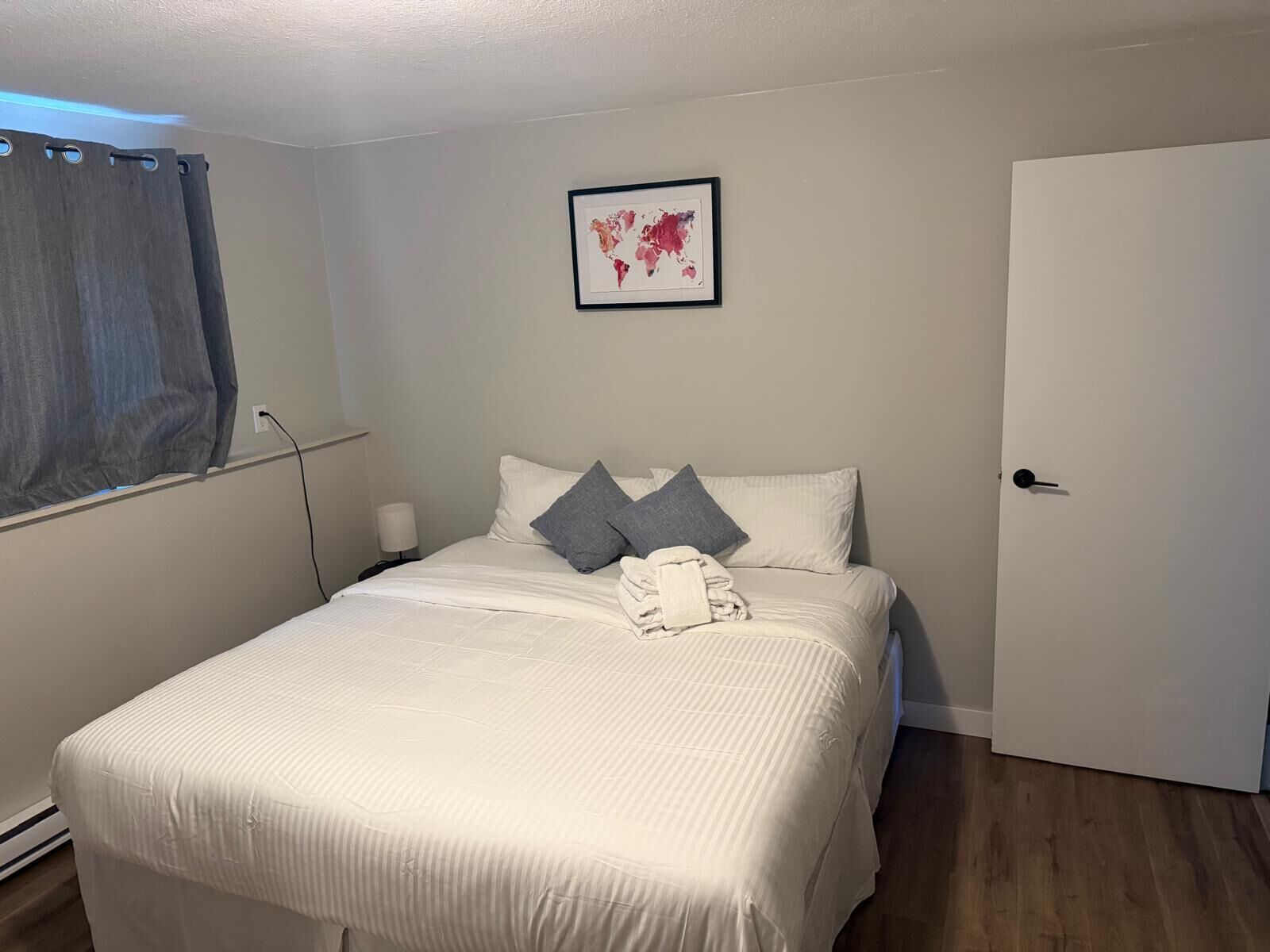 Una cama bien hecha con almohadas y una toalla doblada, un mapa mundial enmarcado en la pared, una lámpara de noche y una puerta a la derecha.