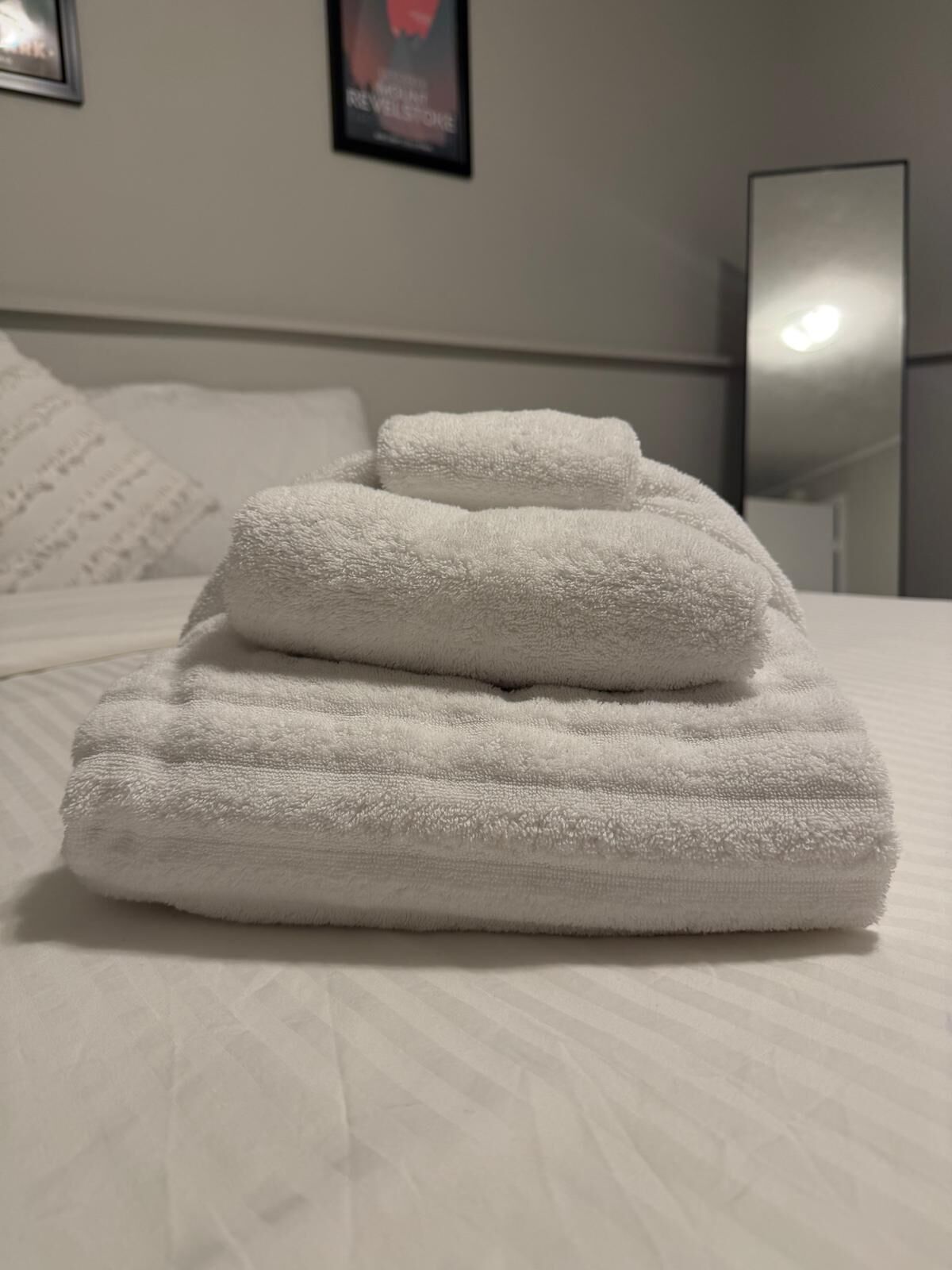 Een keurig opgevouwen witte handdoek op een bed met een wit kussen en een ingelijst foto aan de muur.