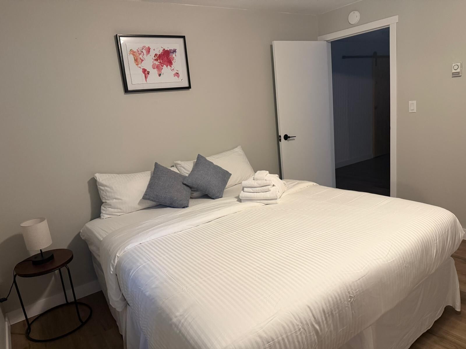 A neatly made bed with white bedding and gray pillows, a framed world map on the wall, and a closed door to the right.