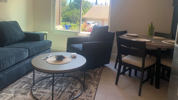 A living room with a sofa, a round coffee table, a dining table with chairs, and a window with a view of trees and buildings. - Valle verde get-away in upscale neiborhood! (Poway)