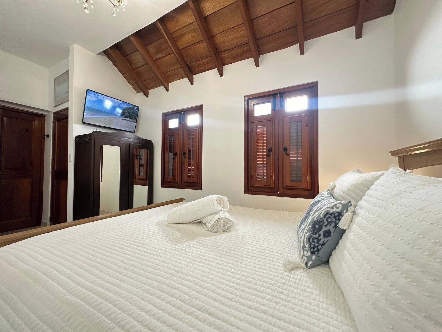 A bedroom with a large bed, wooden ceiling beams, a television on the wall, and two windows with shutters.