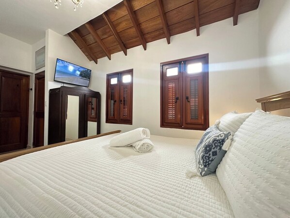 A bedroom with a large bed, wooden ceiling beams, a television on the wall, and two windows with shutters.