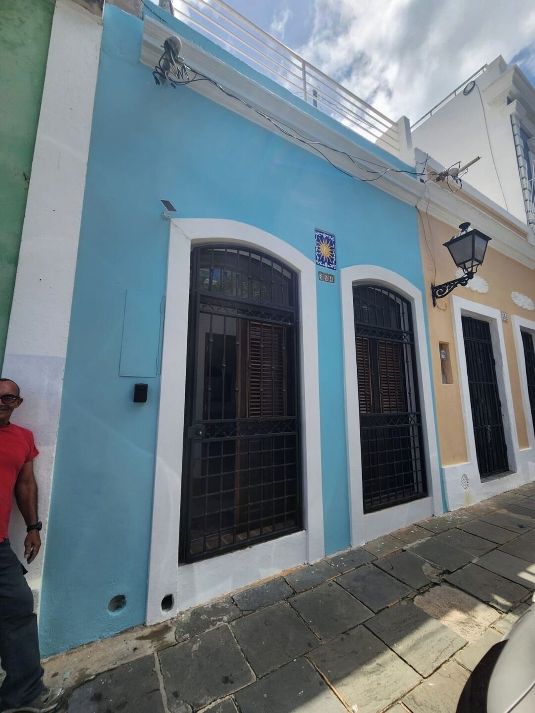 Un edificio con fachada azul y blanca, con grandes ventanas arqueadas con rejas de metal, un panel solar en el techo y una farola.