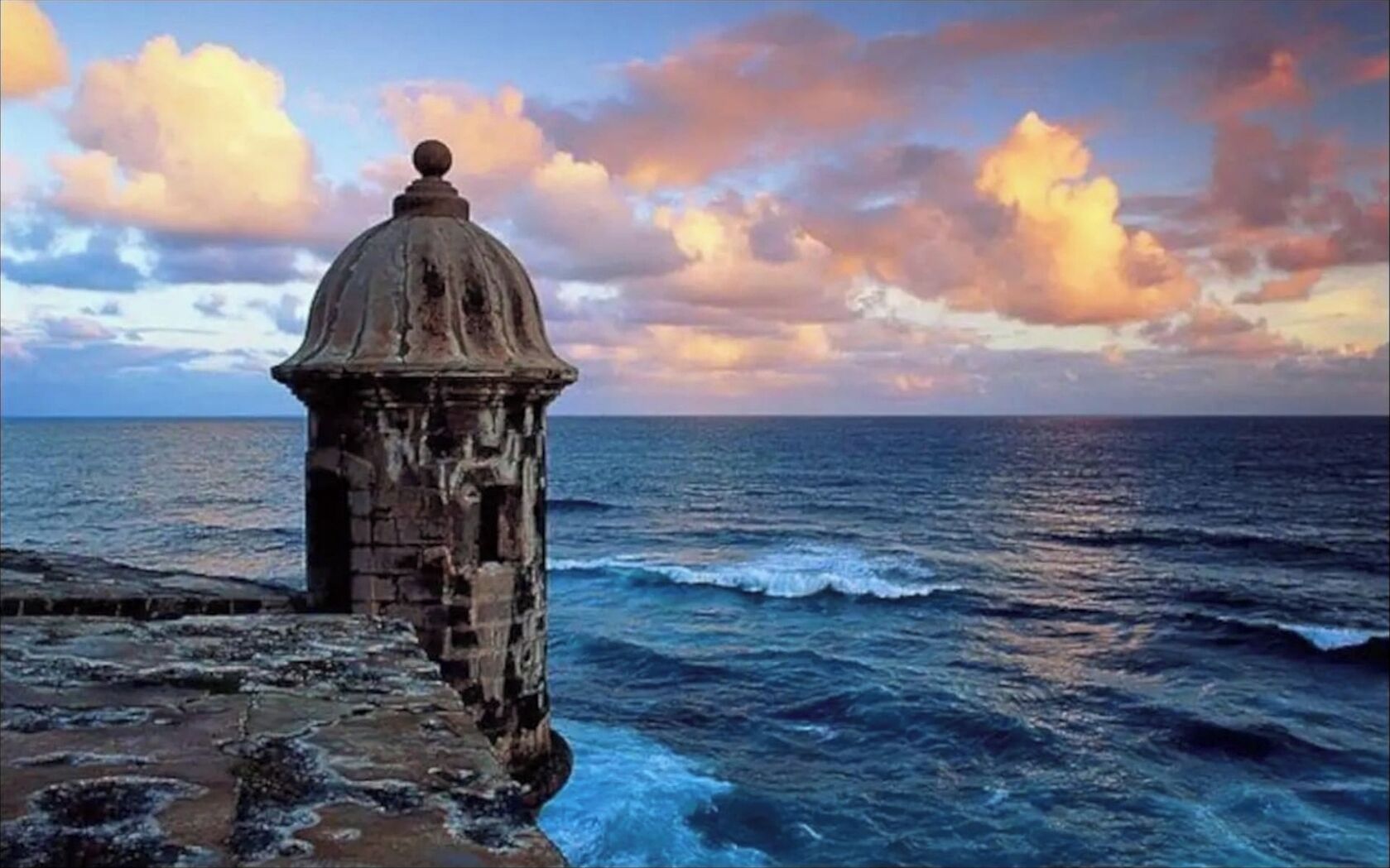 A coastal structure with a dome and a stone base, overlooking the ocean with waves and a sunset sky.
