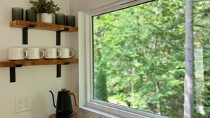 Una cocina con estantes de madera, una cafetera y una ventana con vista al bosque.