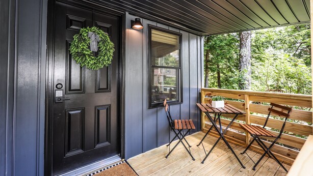 Una puerta de madera oscura con un adorno de guirnalda, una ventana con vistas a árboles y una terraza de madera con dos sillas y una mesa.