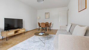 Ein modernes Wohnzimmer mit Flachbildfernseher, einer Holz-Unterhaltungsecke, rundem Couchtisch, beiger Couch und einem Essbereich mit Stühlen und einer Lampe.