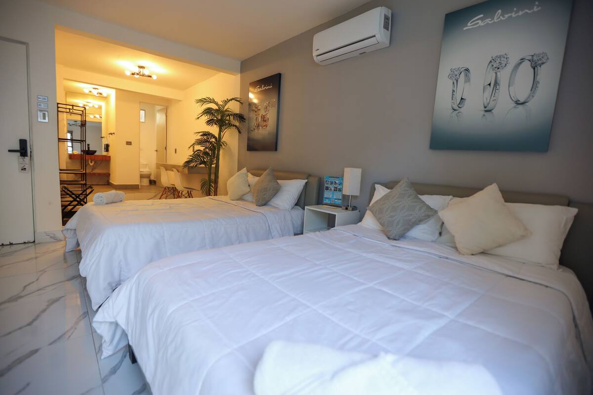 Habitación de hotel con dos camas, un baño, un pasillo y un aire acondicionado instalado en la pared.