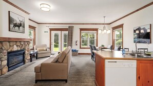 A spacious living room with a stone fireplace, a sofa, a dining area with a kitchen counter, and a large window providing a view of the outdoors.