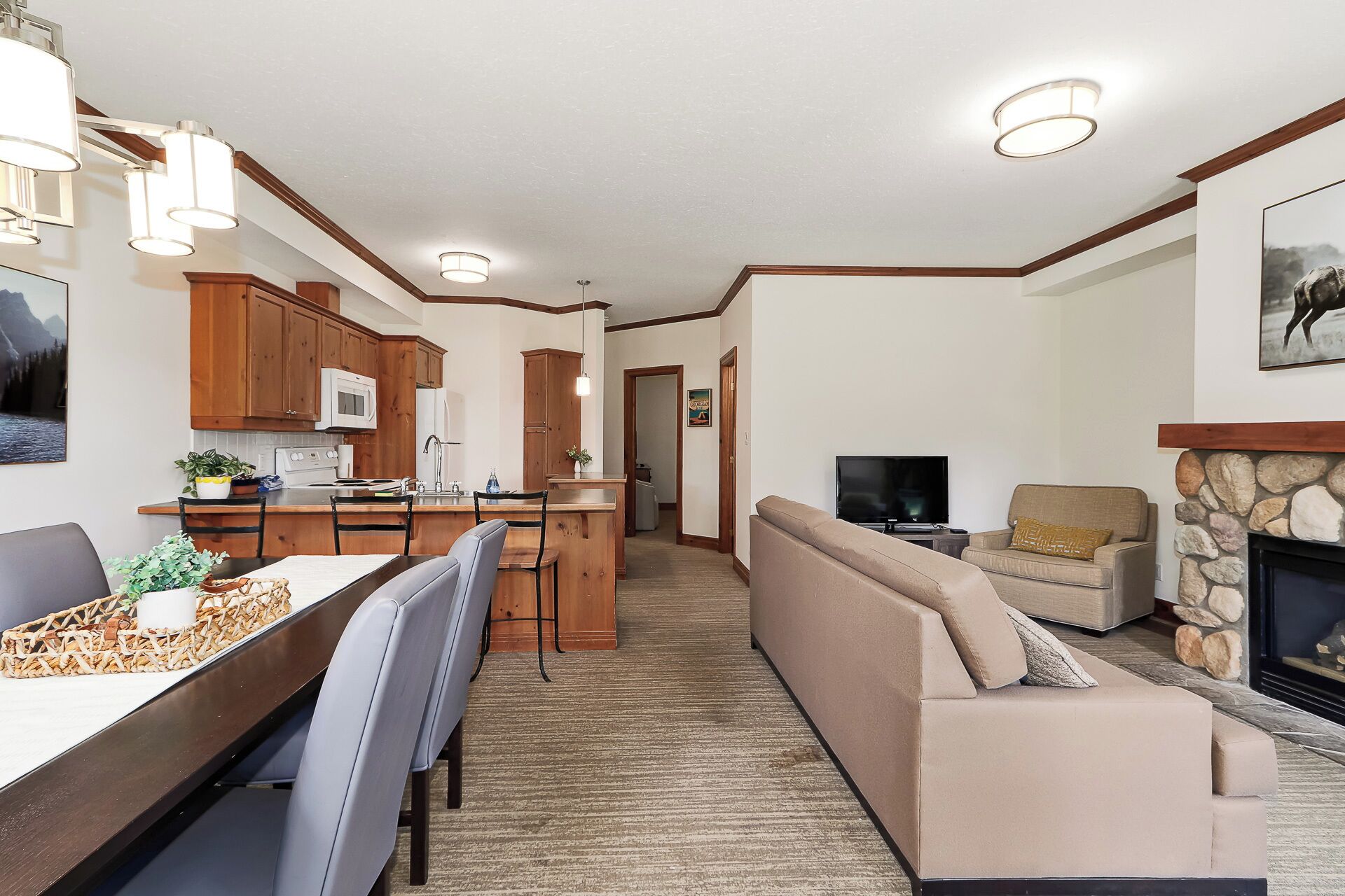 A modern kitchen with wooden cabinets, a dining area with a long table and chairs, a living room with a sofa and a fireplace, and a TV in the corner.