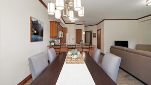 A dining area with a long table, chairs, a chandelier, a kitchen in the background, and a living room with a sofa.