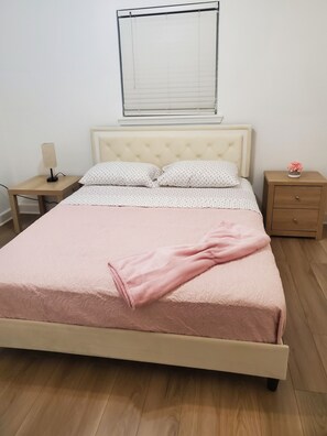 Un letto con una coperta rosa, un comodino con una lampada, un altro comodino con un fiore e una finestra con persiane.