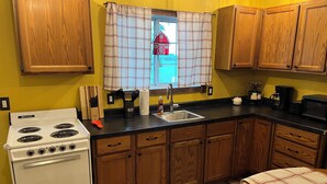 A kitchen with wooden cabinets, a white stove, a black countertop, and a window with plaid curtains. - The Little Red Barn (Cedar Falls)
