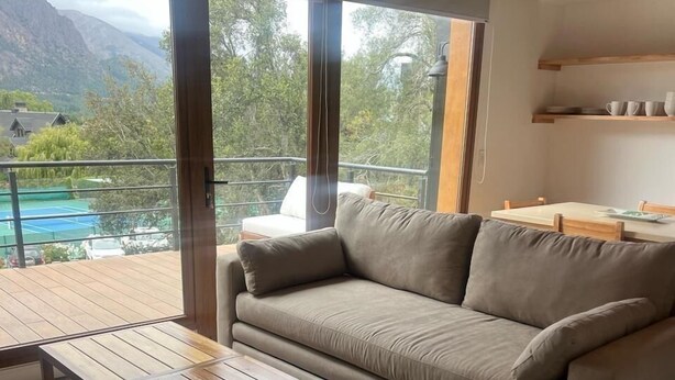 Un salon moderne avec un canapé beige, une table basse en bois et une vue sur un court de tennis et des montagnes, visible à travers de grandes fenêtres.