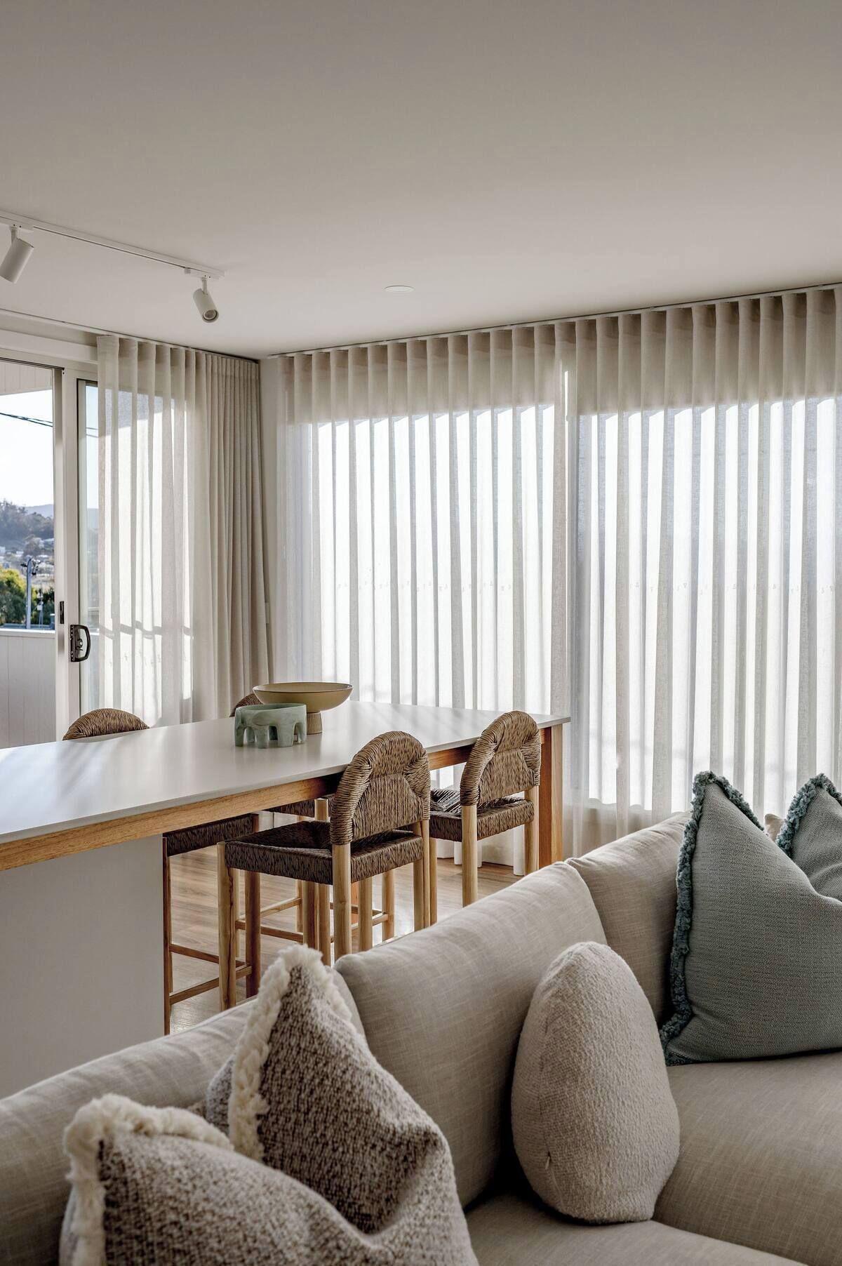 A modern living room with a wooden dining table, wicker chairs, and a beige sofa with throw pillows.