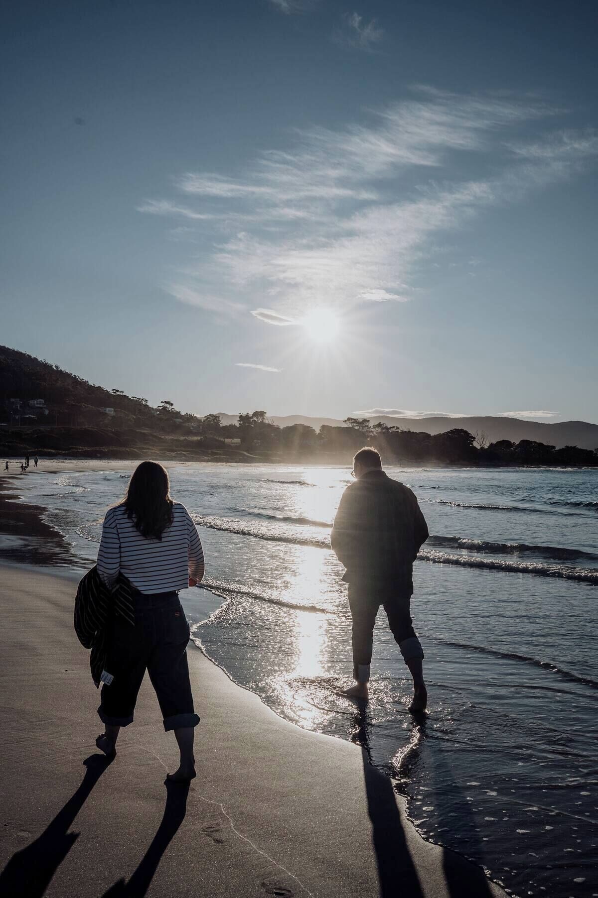 Twee mensen wandelen over een strand, met een ondergaande zon op de achtergrond.