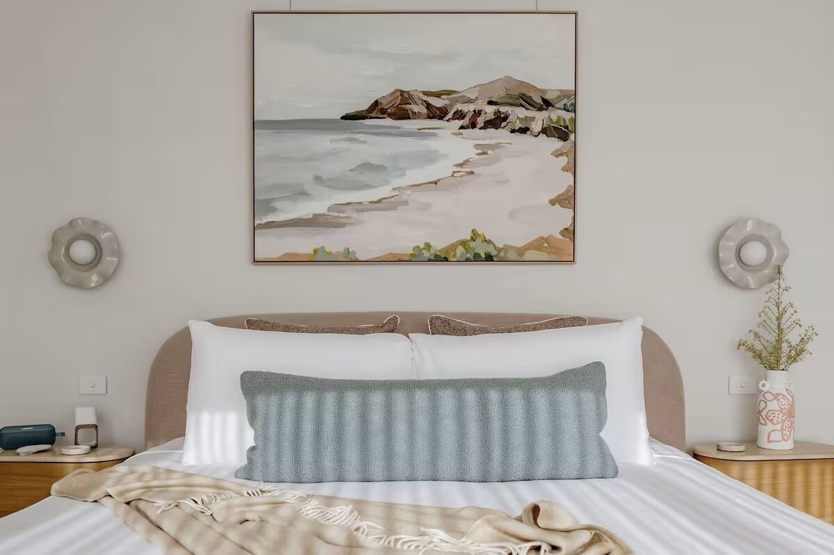 Ein ordentlich bezogenes Bett mit Kissen, ein Nachttisch mit Lampe und ein gerahmtes Bild einer Strandansicht an der Wand.