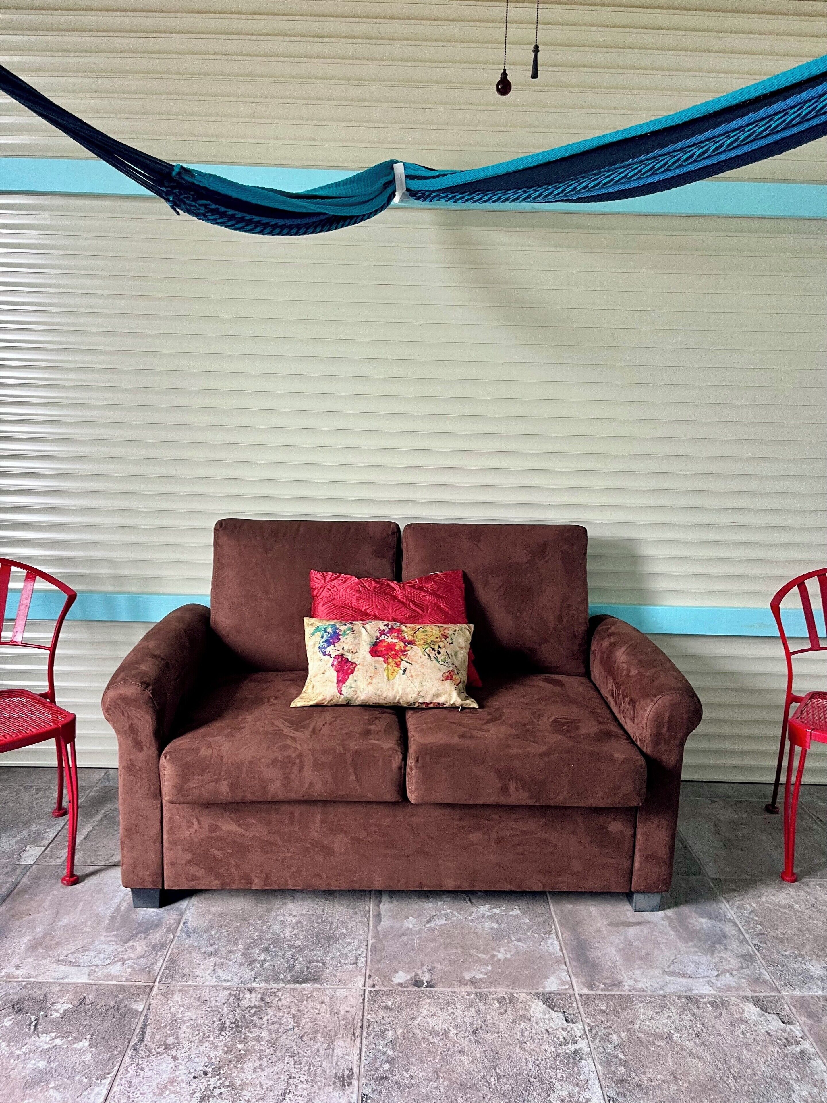 A brown sofa with floral and red cushions, two red chairs, and a blue hammock against a white wall with a blue stripe.