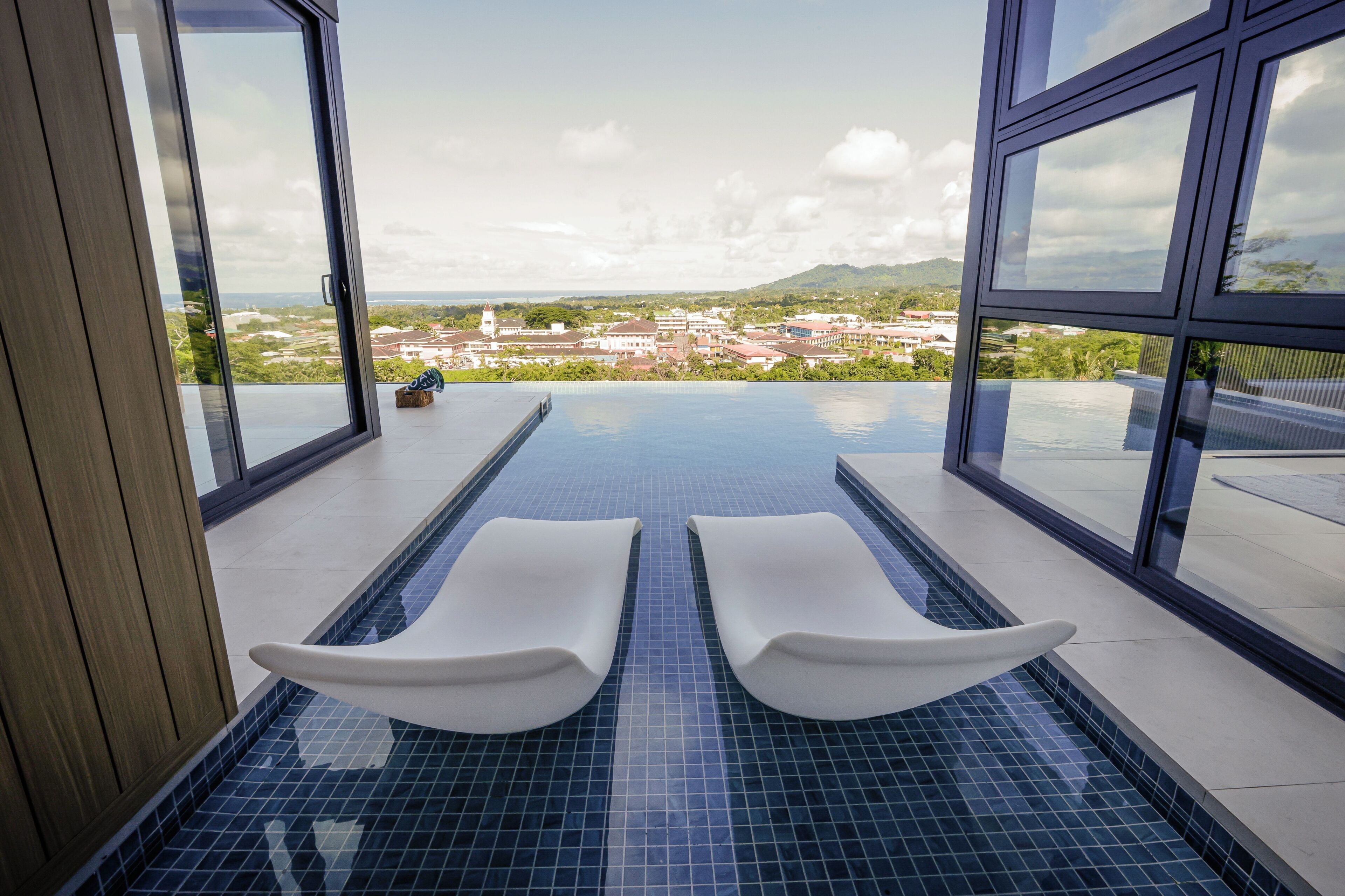 A modern indoor pool with two white loungers, large windows offering a view of a cityscape and mountains.