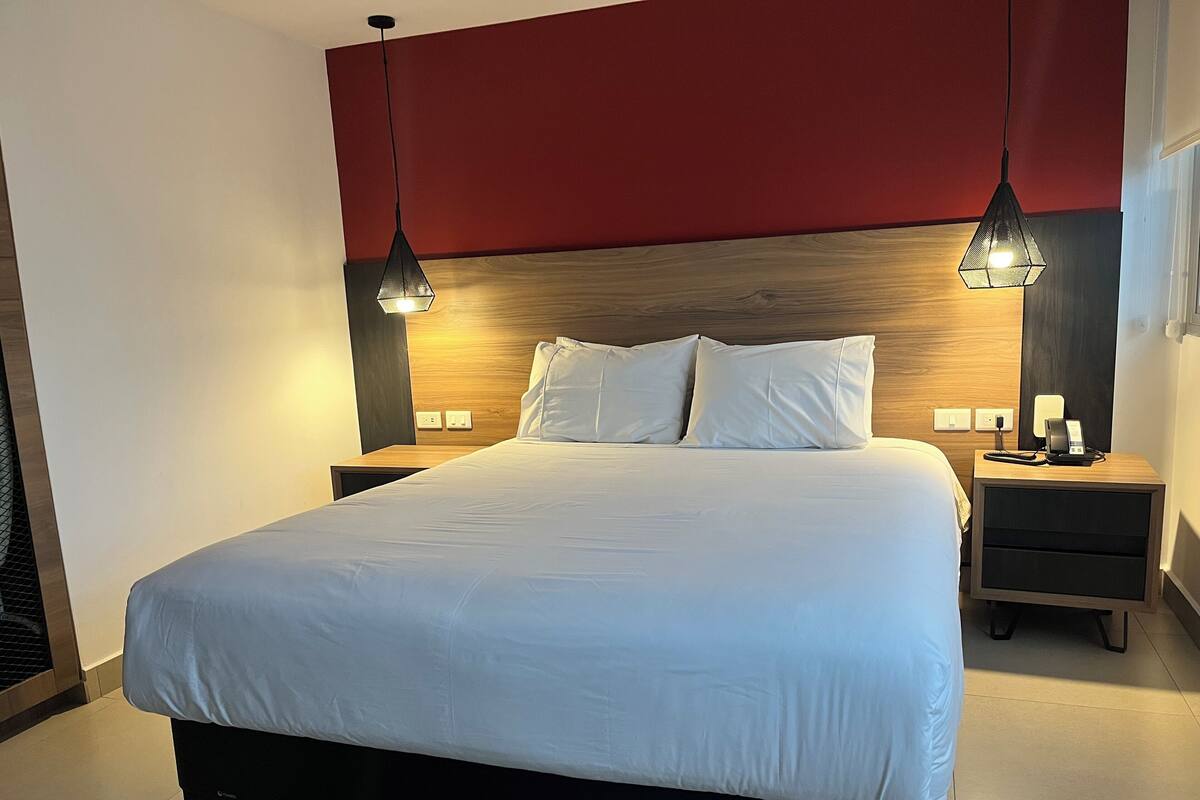 A modern hotel room with a large bed, wooden headboard, two bedside tables with lamps, and a red accent wall.