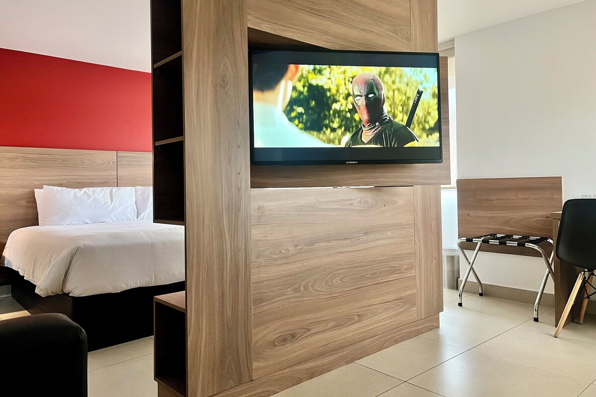 A hotel room with a bed, a TV mounted on a wooden wall, a desk, and a chair.