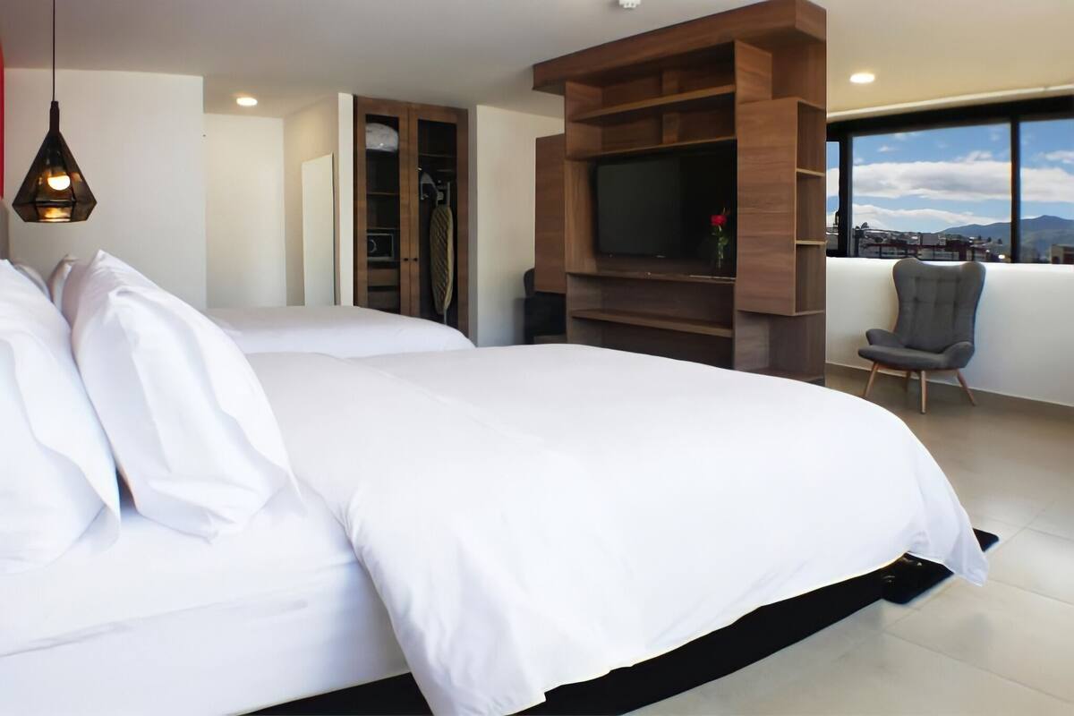 A modern hotel room with a large bed, a TV mounted on a wooden wall, a chair, and a view of the city through large windows.