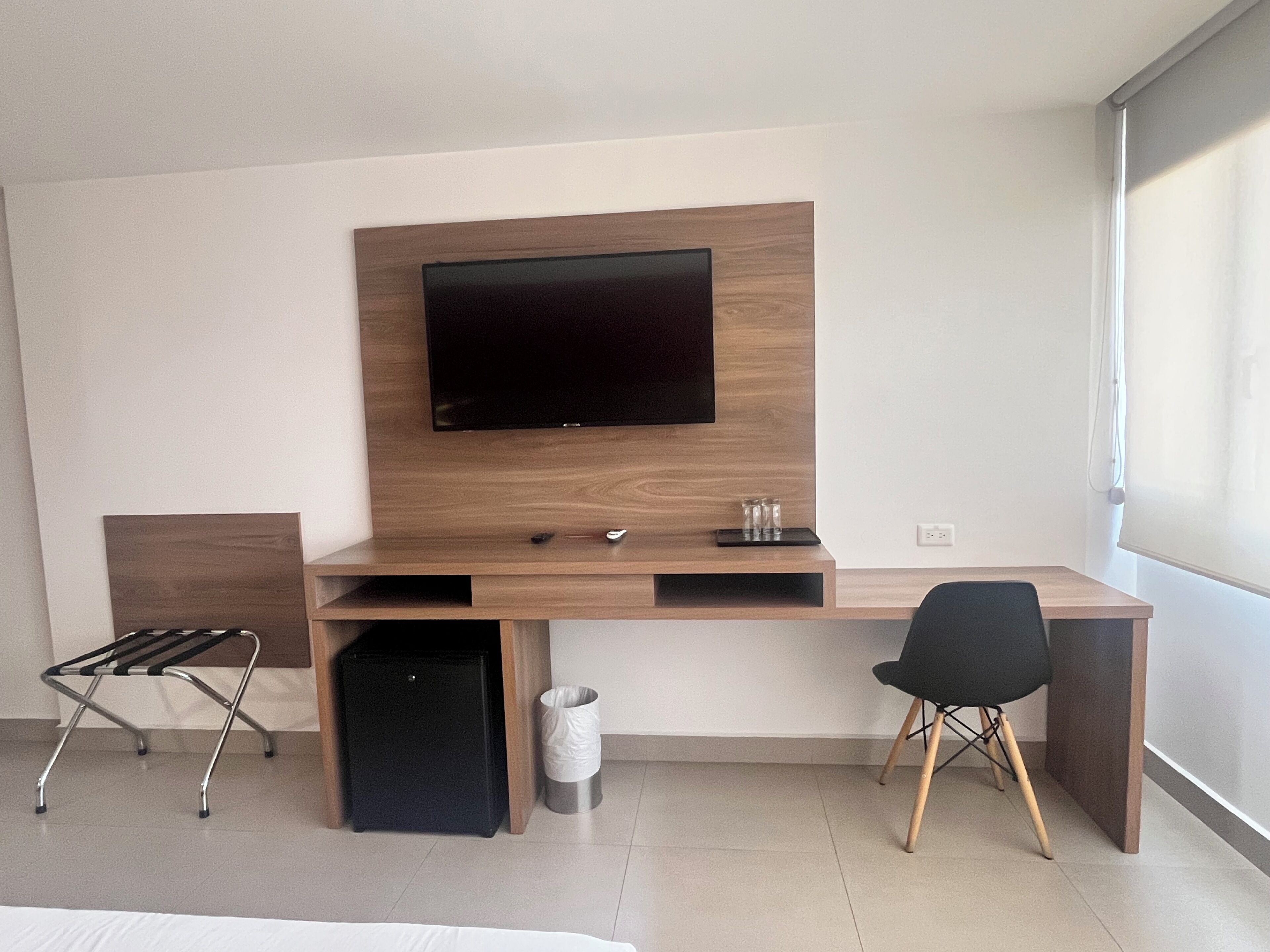 A modern hotel room with a wooden desk, a flat-screen TV, a black chair, a small black speaker, and a waste bin.