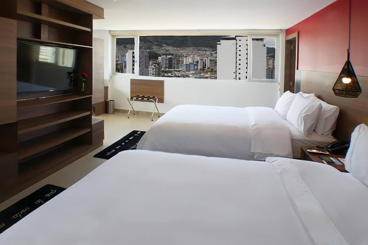 A hotel room with two beds, a TV, a chair, a window with a city view, and a hanging light fixture.