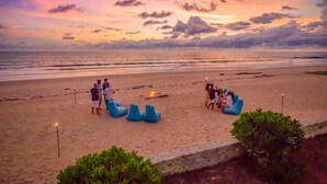 A beach scene with people gathered around a fire pit, surrounded by blue lounge chairs.