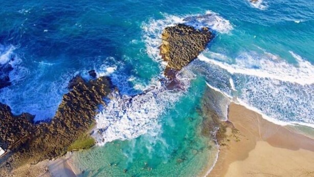 Vista aérea de una playa con aguas turquesas cristalinas, formaciones rocosas y orillas arenosas.
