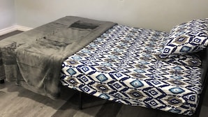 Una habitación pequeña con cama individual, una colcha estampada, una manta gris y una silla negra.
