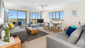 A spacious living room with a large TV, dining area, and a balcony with a view.