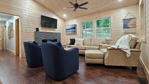 A cozy living room with a stone fireplace, a ceiling fan, and a large window offering a view of the outdoors.