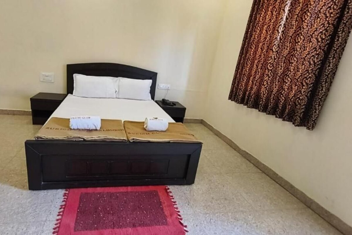 Une chambre d’hôtel avec un lit simple, une table de chevet, un tapis rouge et un rideau à motifs.