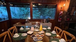 Taula de dinar posta amb plats, coberts i una safata de fruita, envoltada de cadires, amb vista a arbres a través de grans finestres.