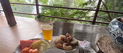 A table with a cup of coffee, a glass of juice, and a plate of food including watermelon, lemon, and pastries, set on a wooden deck with a view of trees.