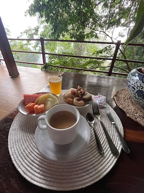 A table with a cup of coffee, a glass of juice, and a plate of food including watermelon, lemon, and pastries, set on a wooden deck with a view of trees.