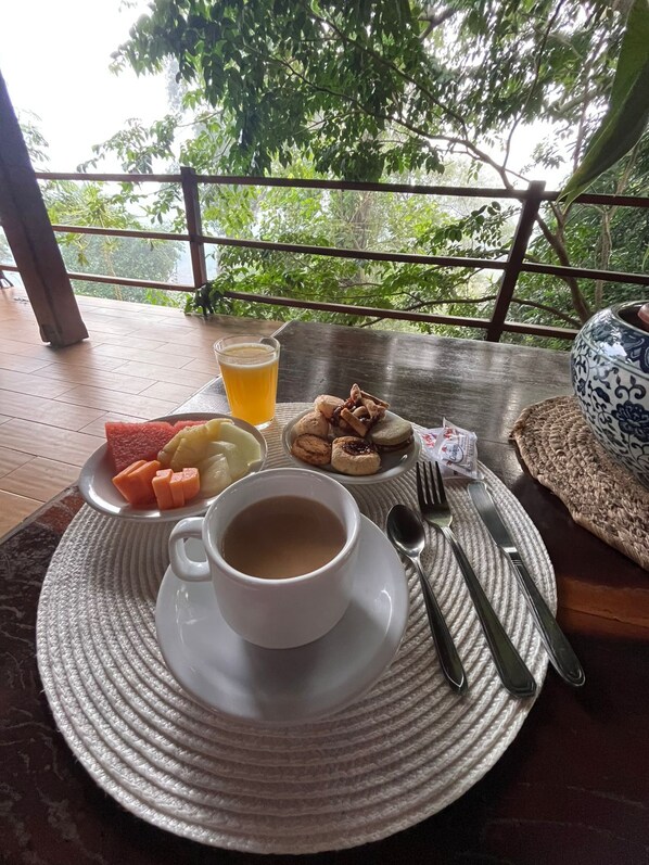 A table with a cup of coffee, a glass of juice, and a plate of food including watermelon, lemon, and pastries, set on a wooden deck with a view of trees. - Botanica The House Experience (Puerto Iguazú)