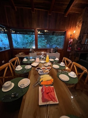Een eettafel is gedekt met borden, bestek en een fruitschaal, omringd door stoelen, met uitzicht op bomen door grote ramen.