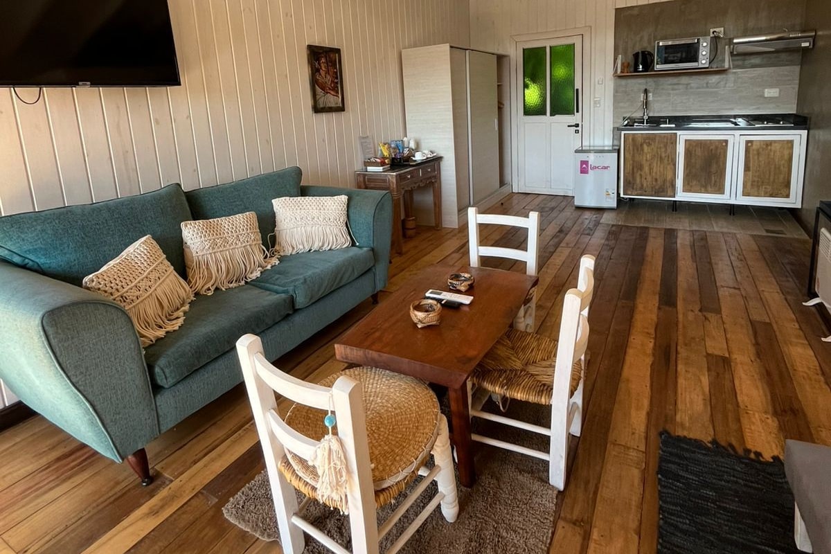 Un vaste espace de vie avec un canapé vert, une table à manger en bois et des chaises blanches. Un téléviseur à écran plat est fixé au mur, et on aperçoit au fond une cuisine équipée d’un four à micro-ondes et d’une cuisinière.