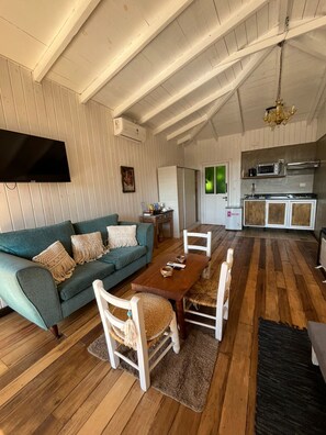 Un'ampia zona giorno con un divano verde, un tavolo da pranzo in legno e sedie bianche. Una TV a schermo piatto è fissata al muro, e sullo sfondo si può vedere una cucina con forno a microonde e piano cottura.