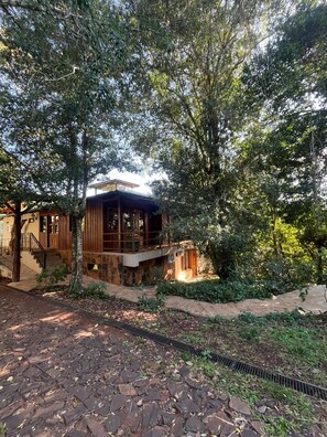 A rustic cabin with a stone foundation, wooden exterior, and a balcony overlooking a garden. - Botanica The House Experience (Puerto Iguazú)