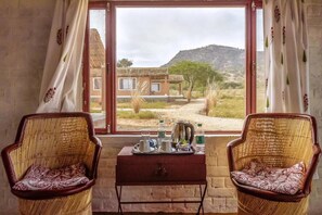 A room with two wicker chairs facing a window overlooking a house and a hilly landscape. A small table with a tray of tea and cups is in front of the window. - Camp Lands End (Pushkar)