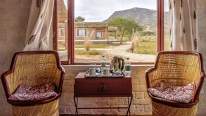 Habitación con dos sillones de mimbre mirando a una ventana con vistas a una casa y un paisaje montañoso. Una mesita con una bandeja de té y tazas se encuentra frente a la ventana.