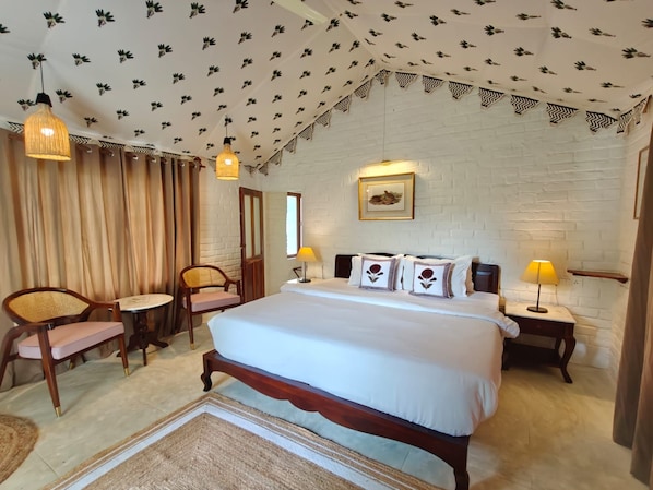 A bedroom with a large bed, two bedside tables, a chair, a small table, and a ceiling with a leaf pattern. - Camp Lands End (Pushkar)