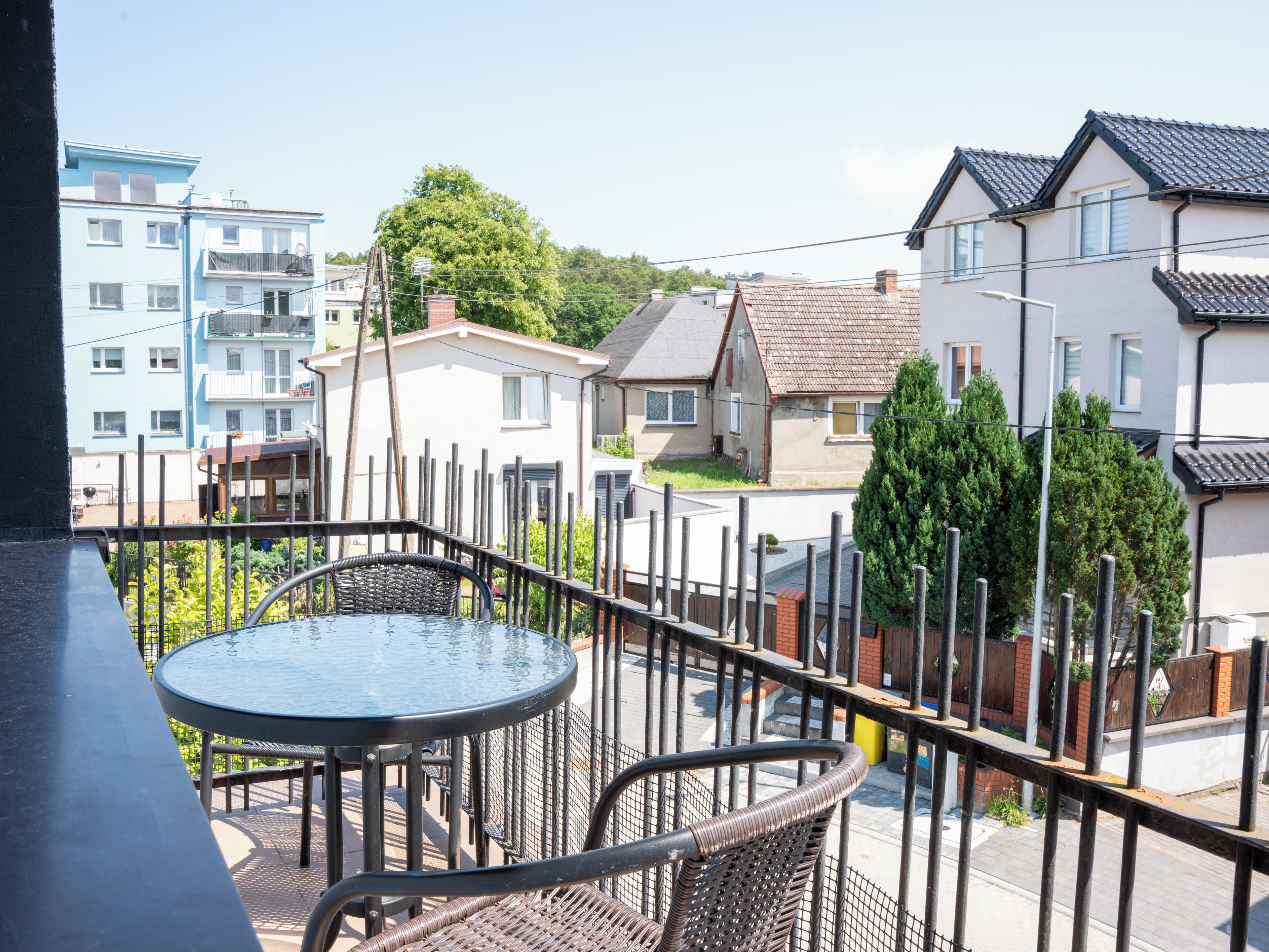 A balcony with a round table and wicker chairs, overlooking a residential area with houses and trees.